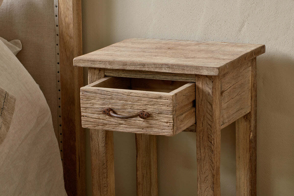 The Ibo Reclaimed Wood Bedside Table by nkuku, featuring antique hardware and an open drawer, stands beside a bed with a neutral headboard and linens.