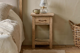 The nkuku Ibo Reclaimed Wood Bedside Table - Natural, featuring a small drawer and lower shelf, stands by a bed with cream bedding. A ceramic mug and glass lamp rest on the table, with a woven basket on the floor and a book on the shelf below.