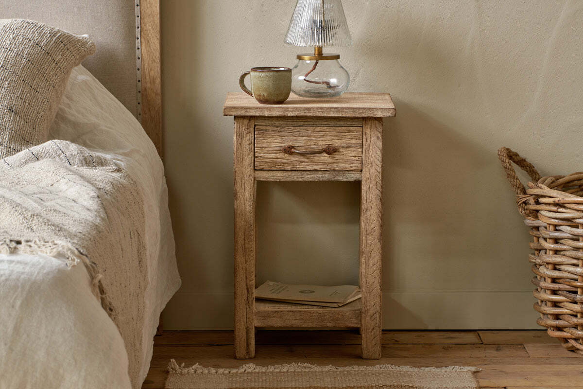 The nkuku Ibo Reclaimed Wood Bedside Table - Natural, featuring a small drawer and lower shelf, stands by a bed with cream bedding. A ceramic mug and glass lamp rest on the table, with a woven basket on the floor and a book on the shelf below.