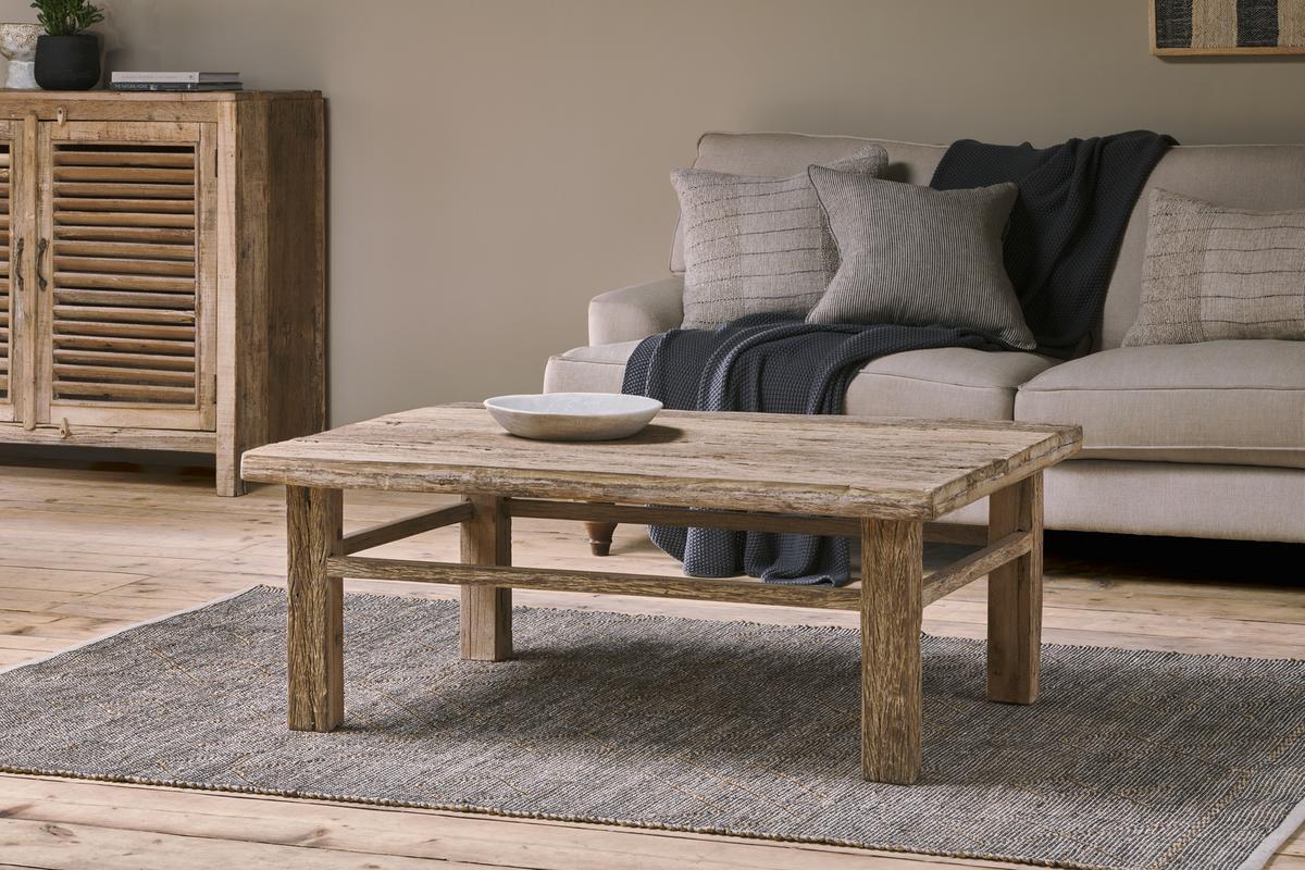 A rustic living room showcases the nkuku Ibo Reclaimed Wood Coffee Table - Natural on a gray rug, topped with a shallow white bowl. Behind it sits a light beige sofa with gray and beige cushions, a dark throw blanket, and a wooden cabinet against the wall.