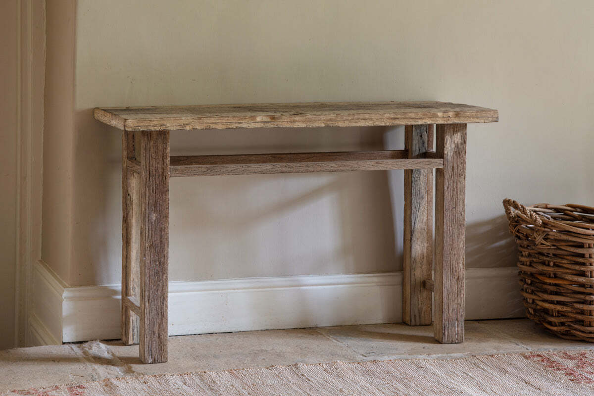 The nkuku Ibo Reclaimed Wood Console Table – Natural stands against a light beige wall, radiating rustic charm. A wicker basket sits to its right.