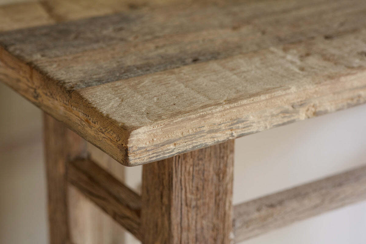 Close-up of the corner of the Ibo Reclaimed Wood Console Table - Natural by nkuku, showcasing its sturdy leg, visible wood grain, and rustic charm crafted from weathered reclaimed wooden panels with an unfinished look.