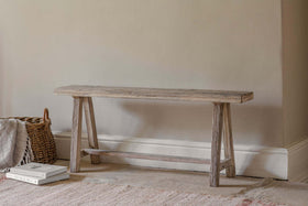 The Ibo Reclaimed Wood Long Bench - Natural by nkuku, featuring simple angled legs, rests against a beige wall. To its left is a woven basket with a folded blanket; two stacked books sit on the light, textured rug.