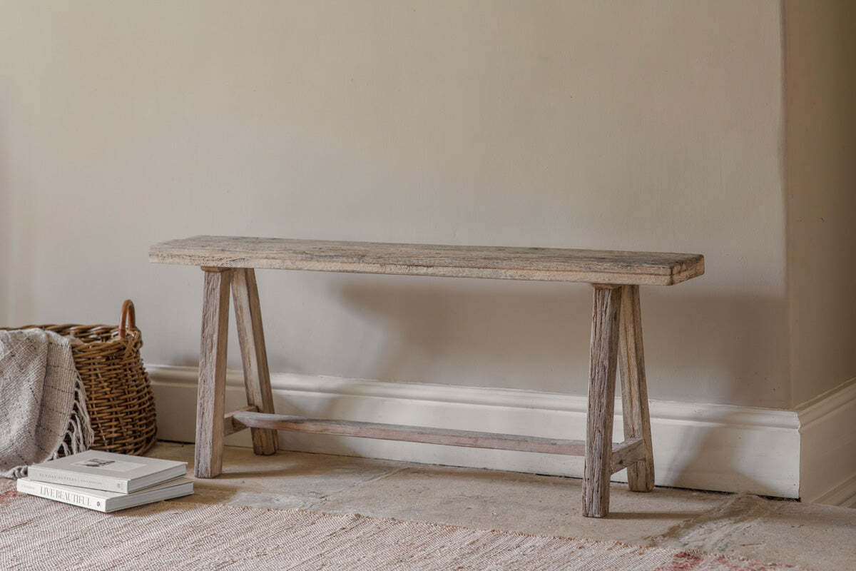 The Ibo Reclaimed Wood Long Bench - Natural by nkuku, featuring simple angled legs, rests against a beige wall. To its left is a woven basket with a folded blanket; two stacked books sit on the light, textured rug.