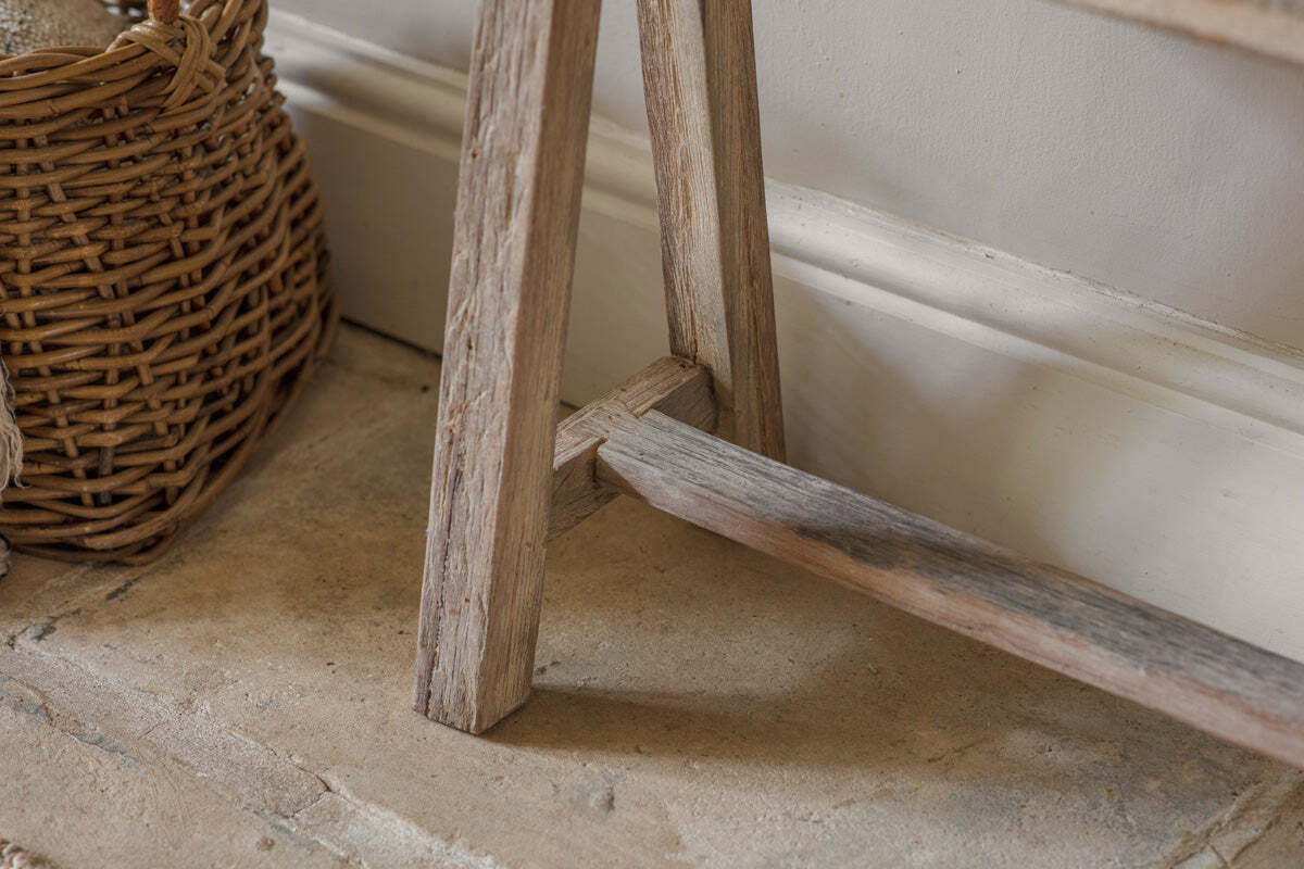 Close-up of the Ibo Reclaimed Wood Long Bench - Natural by nkuku, set on a textured stone floor with a woven wicker basket nearby and a white baseboard against a light wall, creating a cozy, country-inspired atmosphere.