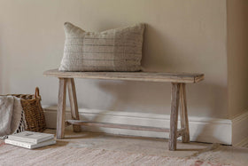 The nkuku Ibo Reclaimed Wood Long Bench - Natural, featuring angled legs, sits by a beige wall. A textured beige cushion tops the bench, while a wicker basket with a blanket and stacked books enhances the cozy, minimalist vibe.