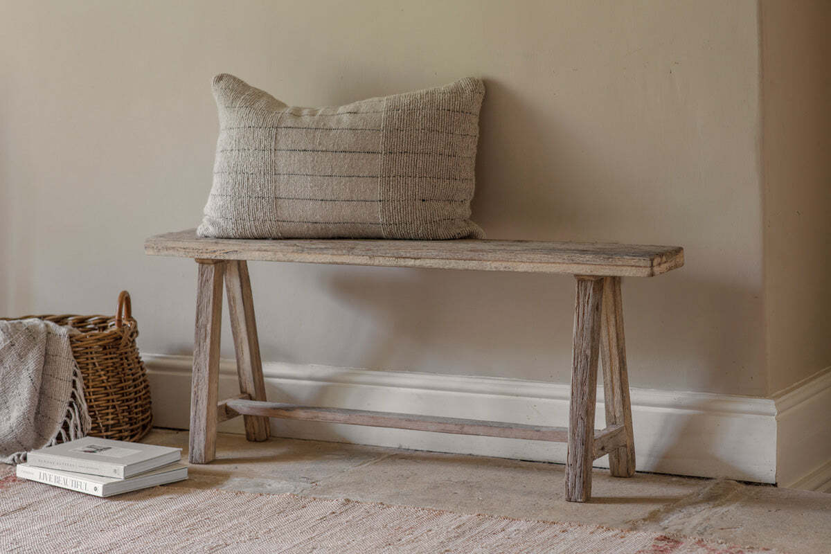 The nkuku Ibo Reclaimed Wood Long Bench - Natural, featuring angled legs, sits by a beige wall. A textured beige cushion tops the bench, while a wicker basket with a blanket and stacked books enhances the cozy, minimalist vibe.