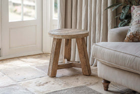 The nkuku Ibo Reclaimed Wood Round Side Table – Natural, with three legs, stands on a stone floor by a beige sofa. Sunlight streams through cream curtains onto this cozy living room accented by a leafy plant and a patterned cushion.