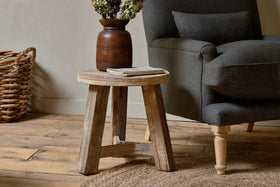 The nkuku Ibo Reclaimed Wood Round Side Table in Natural stands beside a dark gray armchair on a wooden floor, topped with a brown vase of dried flowers and two books, complemented by a woven basket and circular jute rug for a cozy scene.