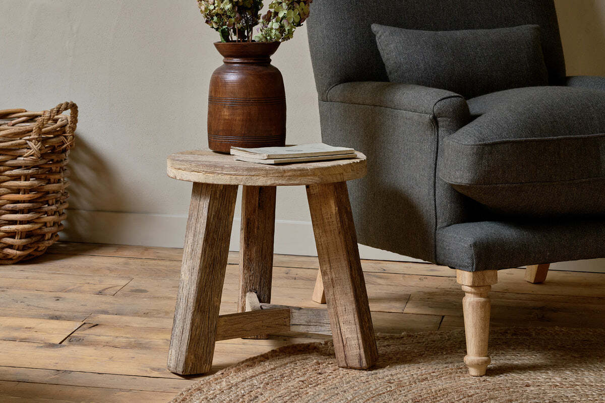 The nkuku Ibo Reclaimed Wood Round Side Table in Natural stands beside a dark gray armchair on a wooden floor, topped with a brown vase of dried flowers and two books, complemented by a woven basket and circular jute rug for a cozy scene.