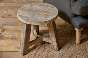 The Ibo Reclaimed Wood Round Side Table - Natural by nkuku features three sturdy legs and a weathered finish, adding cozy, Indian truck wood-inspired charm beside a dark gray sofa and woven jute rug to enhance any natural décor.