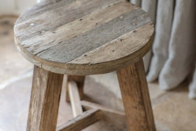 The nkuku Ibo Reclaimed Wood Round Side Table - Natural features a round, weathered wood top with three sturdy legs. A light gray curtain in the background highlights this distinctive piece of reclaimed wood furniture.