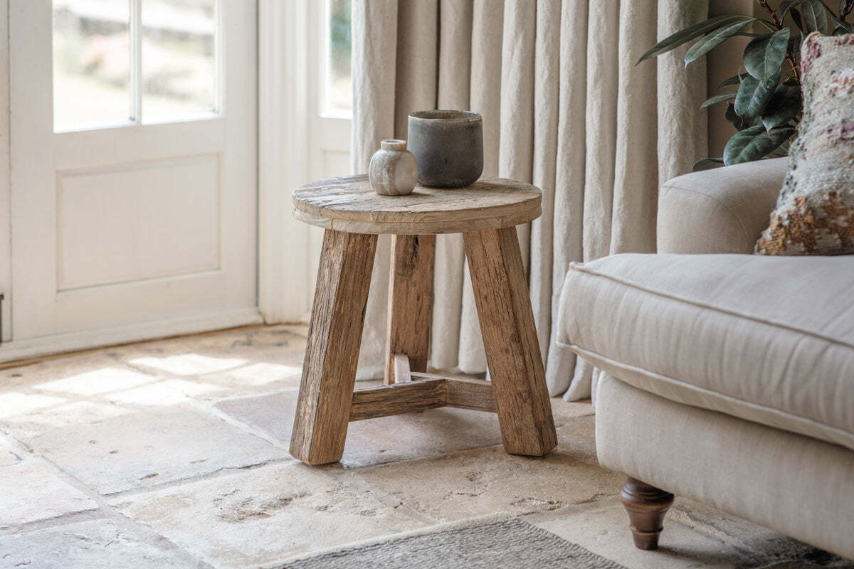 The nkuku Ibo Reclaimed Wood Round Side Table - Natural stands on a stone floor by a beige sofa and cream curtains, topped with white and gray ceramic vases as sunlight streams in through a nearby glass door.