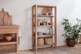The nkuku Ibo Reclaimed Wood Shelf - Natural - Wide holds woven baskets, glass jars, ceramic bowls, plates, and mugs. A potted leafy plant stands beside it. Nearby is a wooden table with cutting boards and bowls on light wood flooring and white walls.
