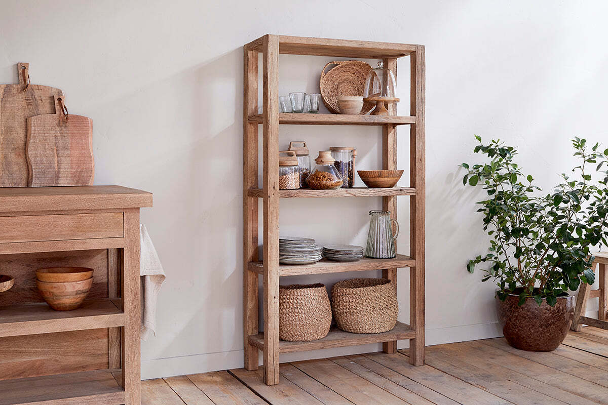 The nkuku Ibo Reclaimed Wood Shelf - Natural - Wide holds woven baskets, glass jars, ceramic bowls, plates, and mugs. A potted leafy plant stands beside it. Nearby is a wooden table with cutting boards and bowls on light wood flooring and white walls.