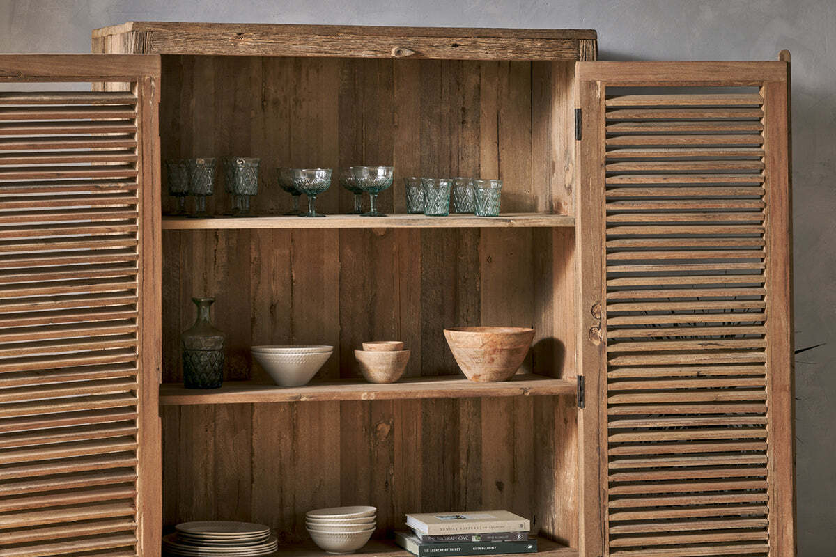 The Ibo Reclaimed Wooden Slatted Cabinet – Natural - Large by nkuku stands open against a light gray wall, revealing three shelves crafted by Indian artisans displaying glassware, bowls, cups, a carafe, and books.