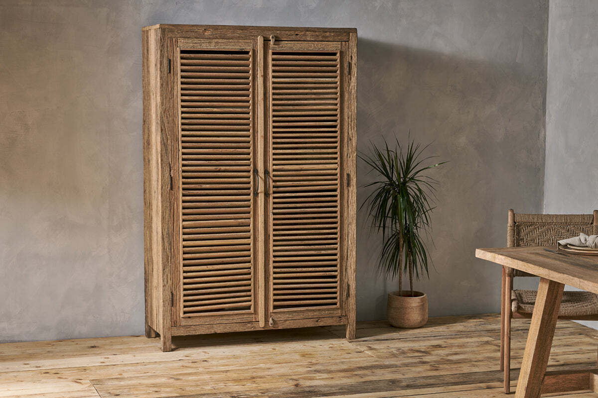 The Ibo Reclaimed Wooden Slatted Cabinet – Natural - Large by nkuku, crafted with Indian artisan skill, stands against a textured gray wall, paired with a potted plant and a wooden table with chair to the right.