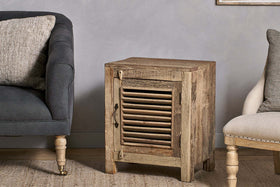 The nkuku Ibo Reclaimed Wooden Slatted Cabinet – Natural - Small, handcrafted by Indian artisans, stands between a dark sofa and a beige chair with wooden legs, resting on a woven rug against a light gray textured wall.