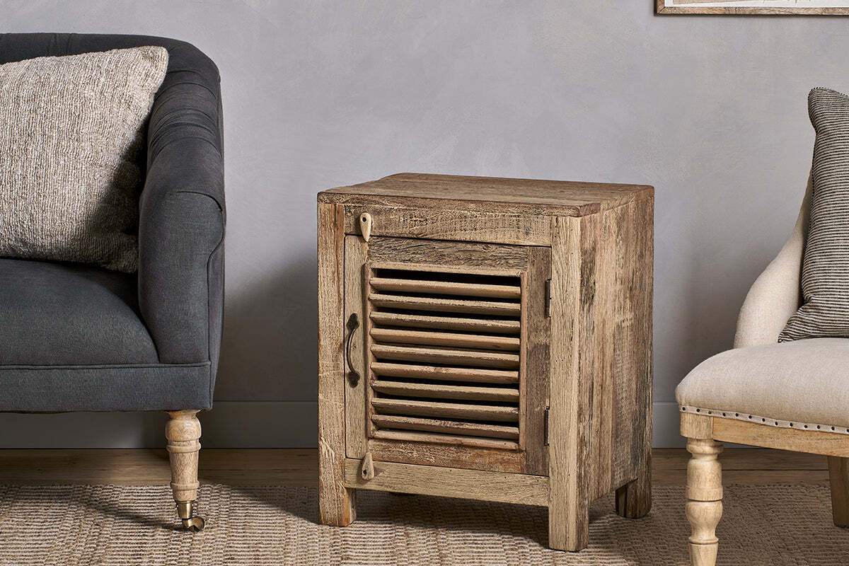 The nkuku Ibo Reclaimed Wooden Slatted Cabinet – Natural - Small, handcrafted by Indian artisans, stands between a dark sofa and a beige chair with wooden legs, resting on a woven rug against a light gray textured wall.