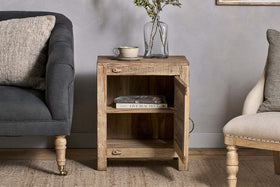 The nkuku Ibo Reclaimed Wooden Slatted Cabinet – Natural - Small sits between a dark gray armchair and a light beige armchair, its open door revealing storage, with a cup, saucer, and glass vase displayed on top.