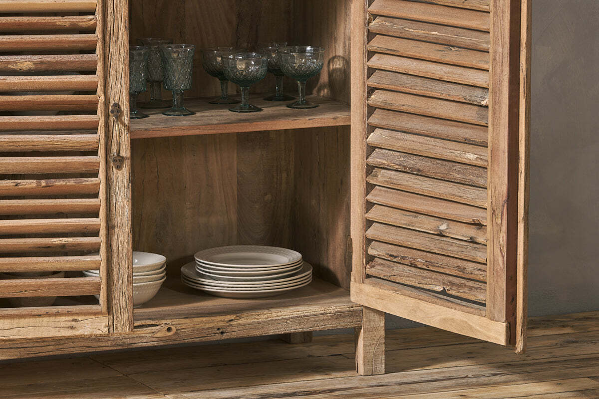 The Ibo Reclaimed Wooden Slatted Sideboard by nkuku stands open, showcasing artisan Indian craftsmanship with interior shelves holding green-tinted glass goblets and white ceramic plates above a wooden floor.