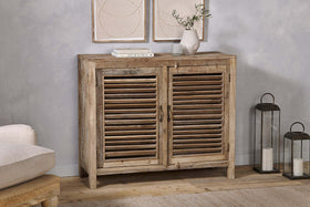 The Ibo Reclaimed Wooden Slatted Sideboard by nkuku features a natural finish and classic Indian craftsmanship. Topped with books, a vase of olive branches, and a glass, it's flanked by two metal lanterns with candles on the right.
