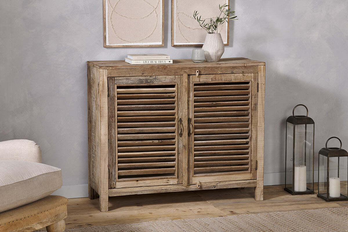 The Ibo Reclaimed Wooden Slatted Sideboard by nkuku features a natural finish and classic Indian craftsmanship. Topped with books, a vase of olive branches, and a glass, it's flanked by two metal lanterns with candles on the right.