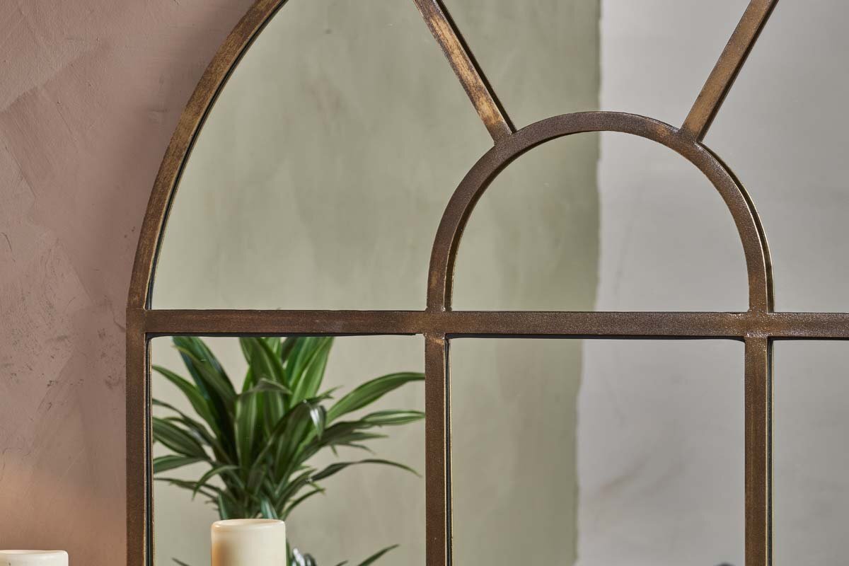 A close-up shows the nkuku Imoma Iron Overmantel Arch Mirror in a brass finish, reflecting a green plant in the lower left pane and bringing a natural touch to its elegant design.