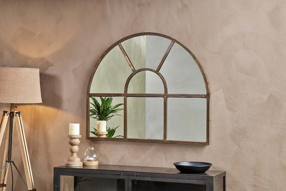 The nkuku Imoma Iron Overmantel Arch Mirror in brass hangs on a beige textured wall above a dark console. Reflected in the mirror are a plant, wooden tripod lamp, two pillar candles on stands, glass cloche, and black bowl.