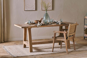 The nkuku Indali Mango Wood Dining Table – Natural is styled with plates, glasses, a clear vase of dried grasses, and woven placemats. Ideal for small spaces, it pairs with a woven-back chair on a textured rug in a softly lit neutral room.
