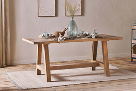 The nkuku Indali Mango Wood Dining Table - Natural, with angled legs, stands on a beige rug in a cozy small space, set with glassware and a vase of dried flowers beneath neutral wall art as sunlight streams through the curtains.