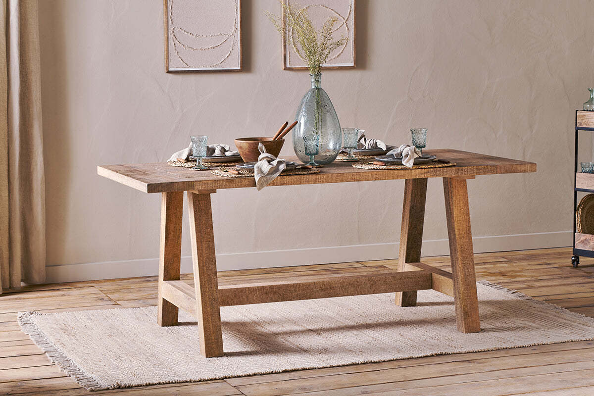 The nkuku Indali Mango Wood Dining Table - Natural, with angled legs, stands on a beige rug in a cozy small space, set with glassware and a vase of dried flowers beneath neutral wall art as sunlight streams through the curtains.
