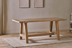 The Indali Mango Wood Dining Table - Natural by nkuku, with angled legs, sits on a beige textured rug in a small space with light wood floors, neutral walls, and two abstract artworks softly lit by natural light.