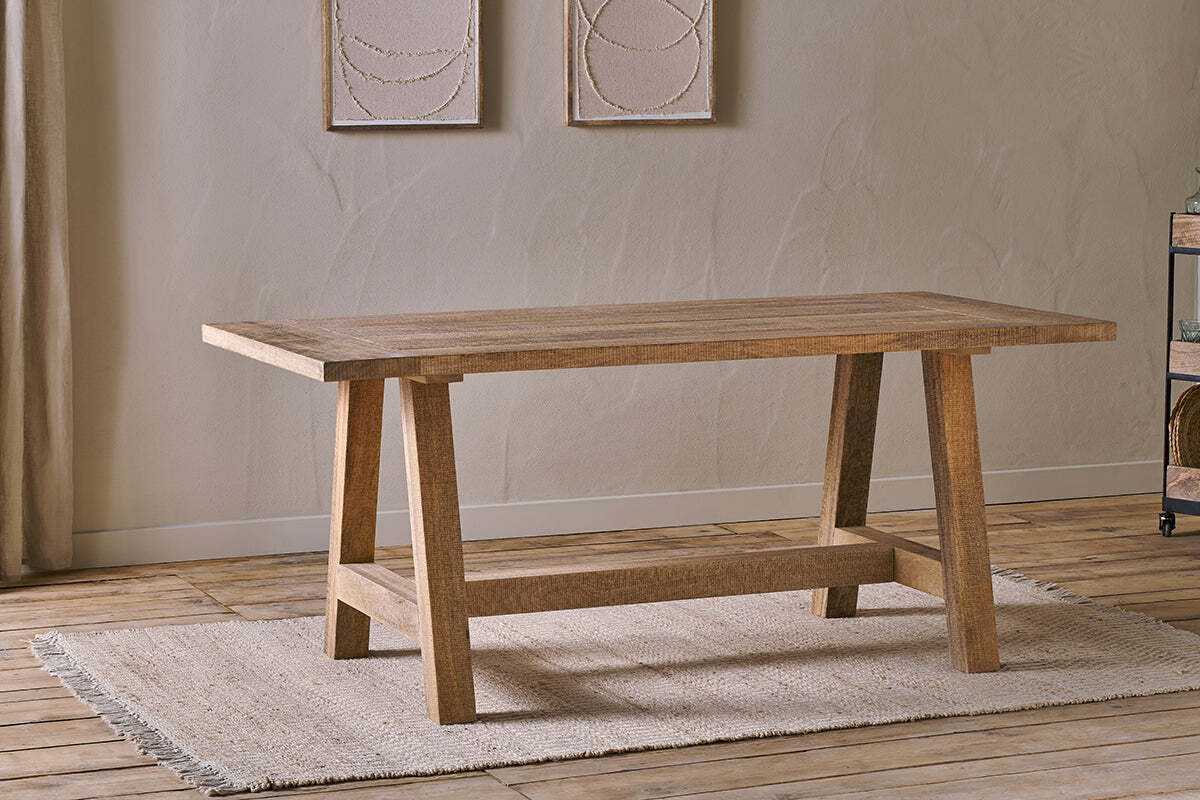 The Indali Mango Wood Dining Table - Natural by nkuku, with angled legs, sits on a beige textured rug in a small space with light wood floors, neutral walls, and two abstract artworks softly lit by natural light.
