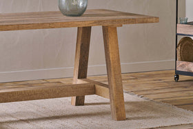 A close-up of the nkuku Indali Mango Wood Dining Table - Natural, featuring angled legs on a textured beige rug. Perfect for small spaces, it displays a rustic finish with a ceramic vase, wooden floor, and metal shelves in the background.