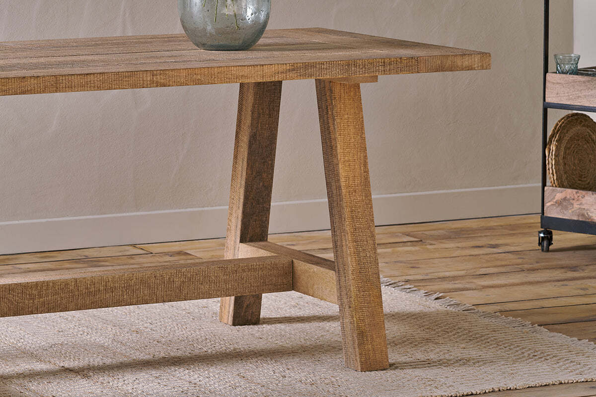 A close-up of the nkuku Indali Mango Wood Dining Table - Natural, featuring angled legs on a textured beige rug. Perfect for small spaces, it displays a rustic finish with a ceramic vase, wooden floor, and metal shelves in the background.