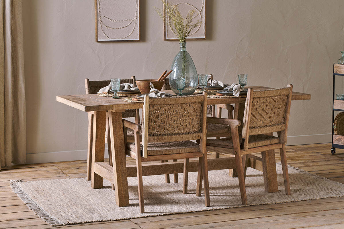 A rustic dining room showcases the nkuku Indali Mango Wood Dining Table - Natural, set for four. Woven chairs, a beige rug, and a large glass vase with dried flowers create an inviting setup ideal for small spaces.