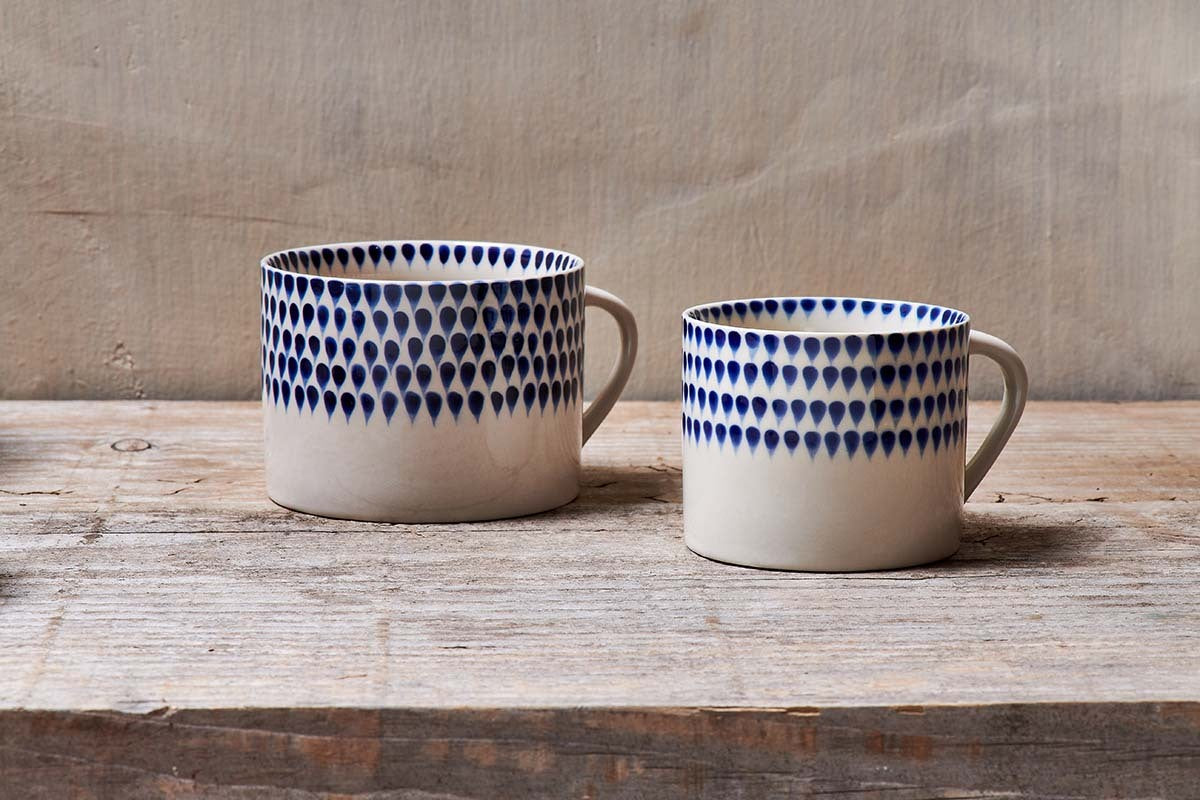 The nkuku Indigo Drop Mug - Cream & Indigo - Small (Set of 2) features hand-painted indigo dots forming a gradient on white ceramic, with both mugs resting on a rustic wooden surface against a neutral backdrop.