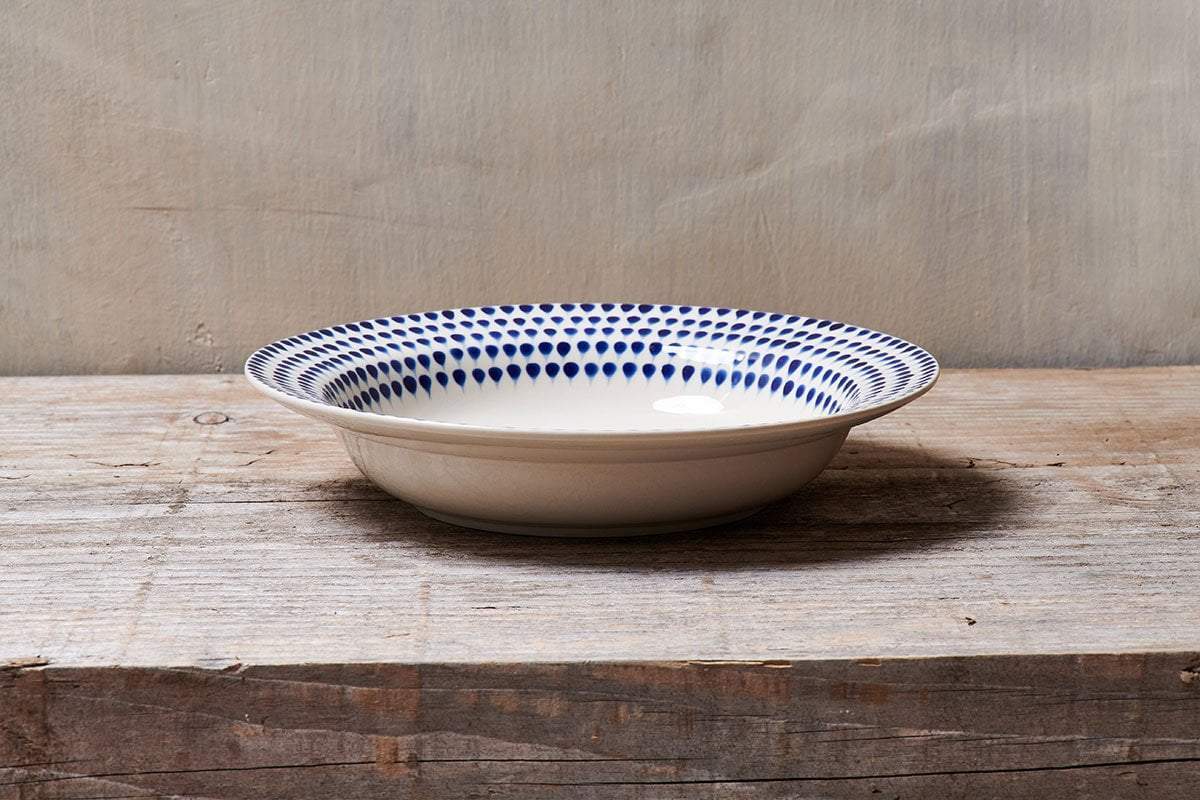 The nkuku Indigo Drop Pasta Bowl - Cream & Indigo features a shallow, round design with a cream base and handpainted blue dot rim, displayed on a rustic wooden surface against a neutral beige wall.