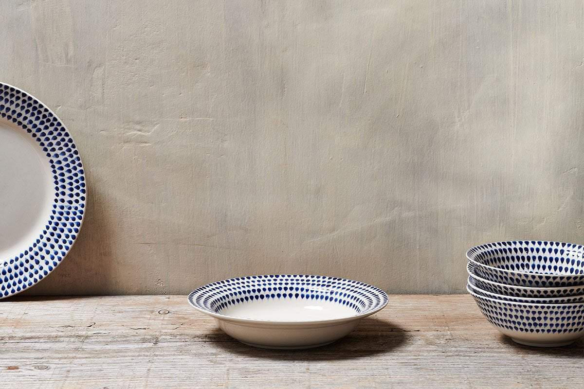 On a rustic wooden surface, the nkuku Indigo Drop Pasta Bowl - Cream & Indigo is displayed alongside handpainted bowls, their blue dotted patterns creating a charming, artisanal look against a plain beige wall.