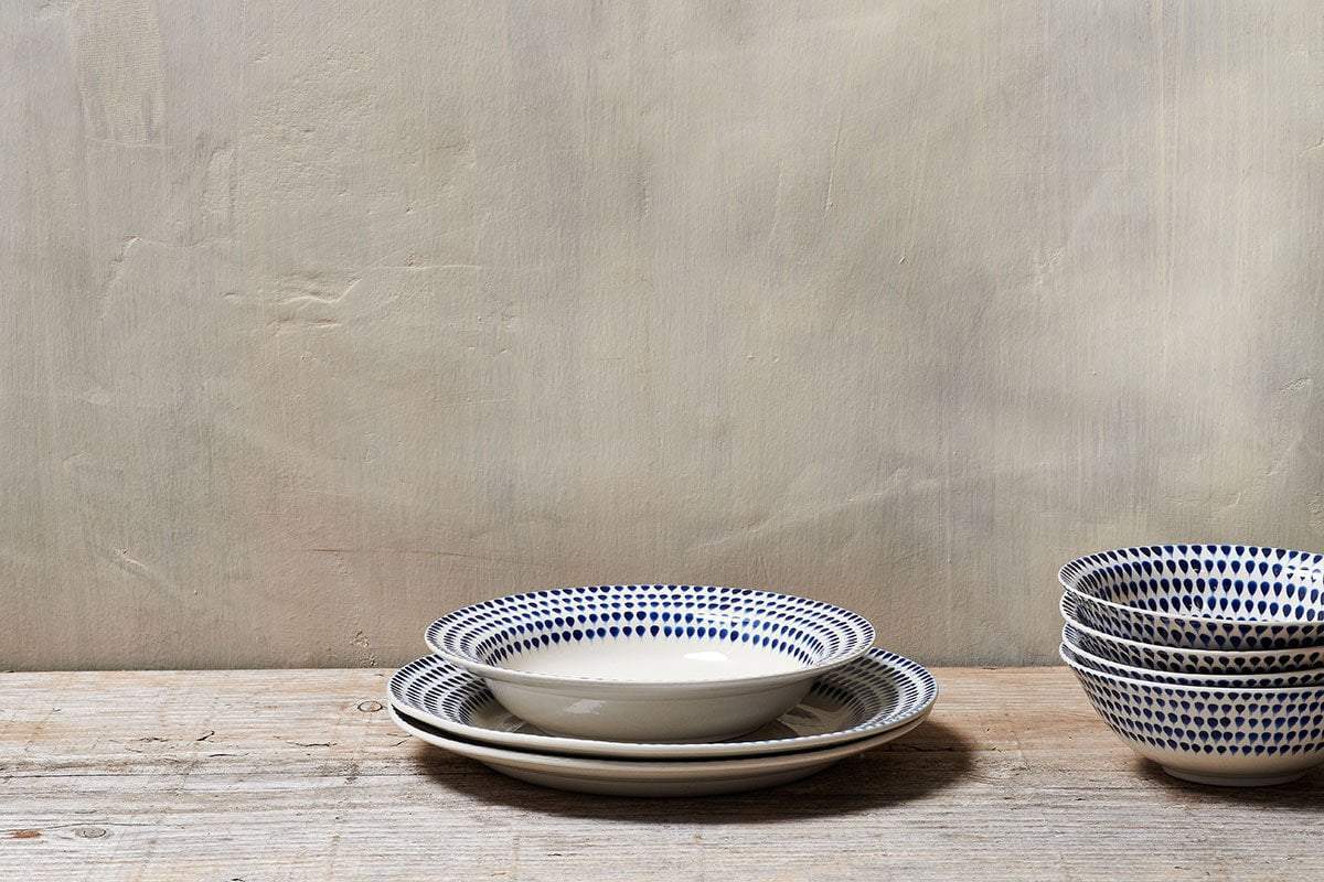 Three handmade ceramic plates with blue dotted patterns are stacked on a rustic wooden surface, alongside four nkuku Indigo Drop Pasta Bowls in Cream & Indigo. A plain beige textured wall forms the minimalist background.