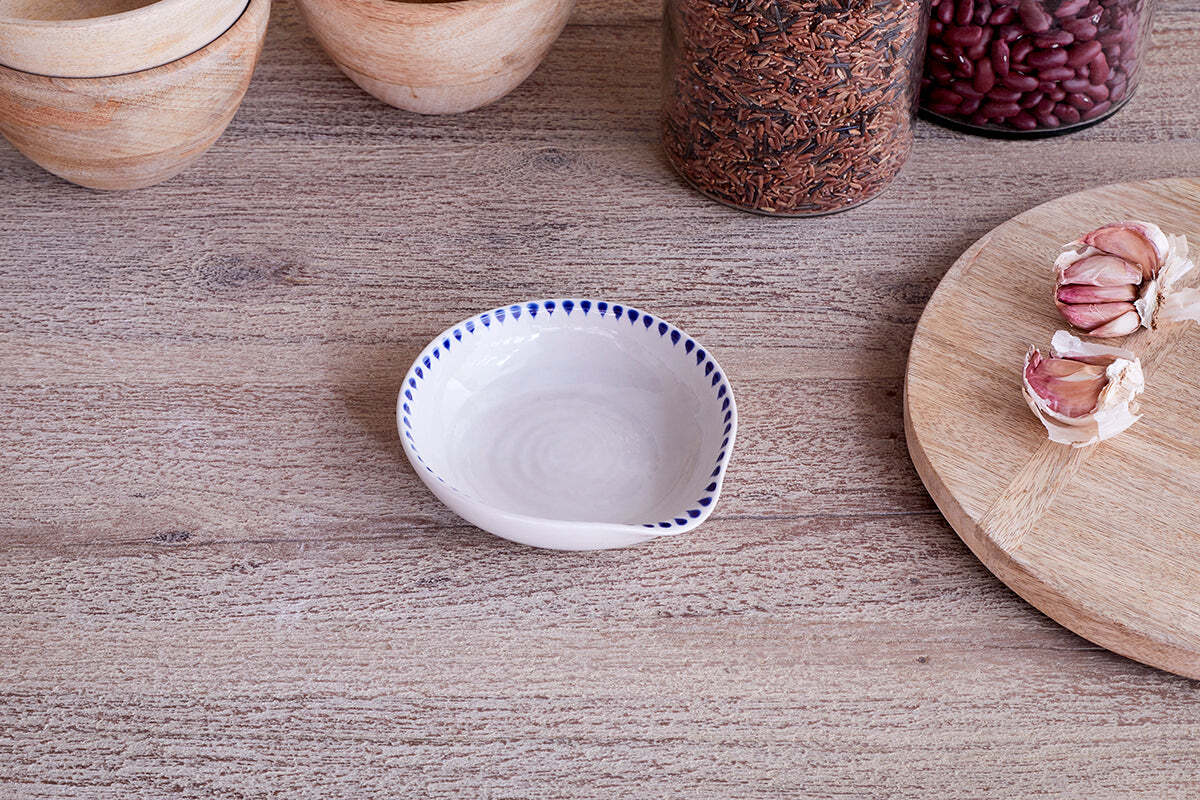 The nkuku Indigo Drop Spoon Rest - Cream & Indigo, featuring hand-painted blue dots, sits on a wooden surface alongside wooden bowls, a jar of rice, red beans, a round board, and garlic bulbs for a rustic kitchen touch.