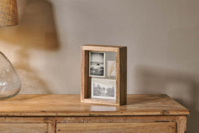 The nkuku Indu Standing Mango Wood Frame - Natural holds two black-and-white photos and text on a rustic dresser, with a glass lamp and beige shade partially visible to the left against a plain light wall.