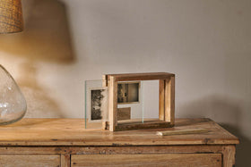The Indu Standing Mango Wood Frame - Natural by nkuku, made from sustainable mango wood, holds two black-and-white photos on a wooden table with a glass lamp. Warm light highlights the frame near a plain beige wall, chalk resting nearby.