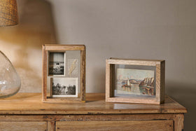 Two Indu Standing Mango Wood Frames by nkuku sit on a wooden surface. The left frame holds three black-and-white photos and a note; the right one displays a colorful riverside painting. A glass lamp is partially visible to the left.