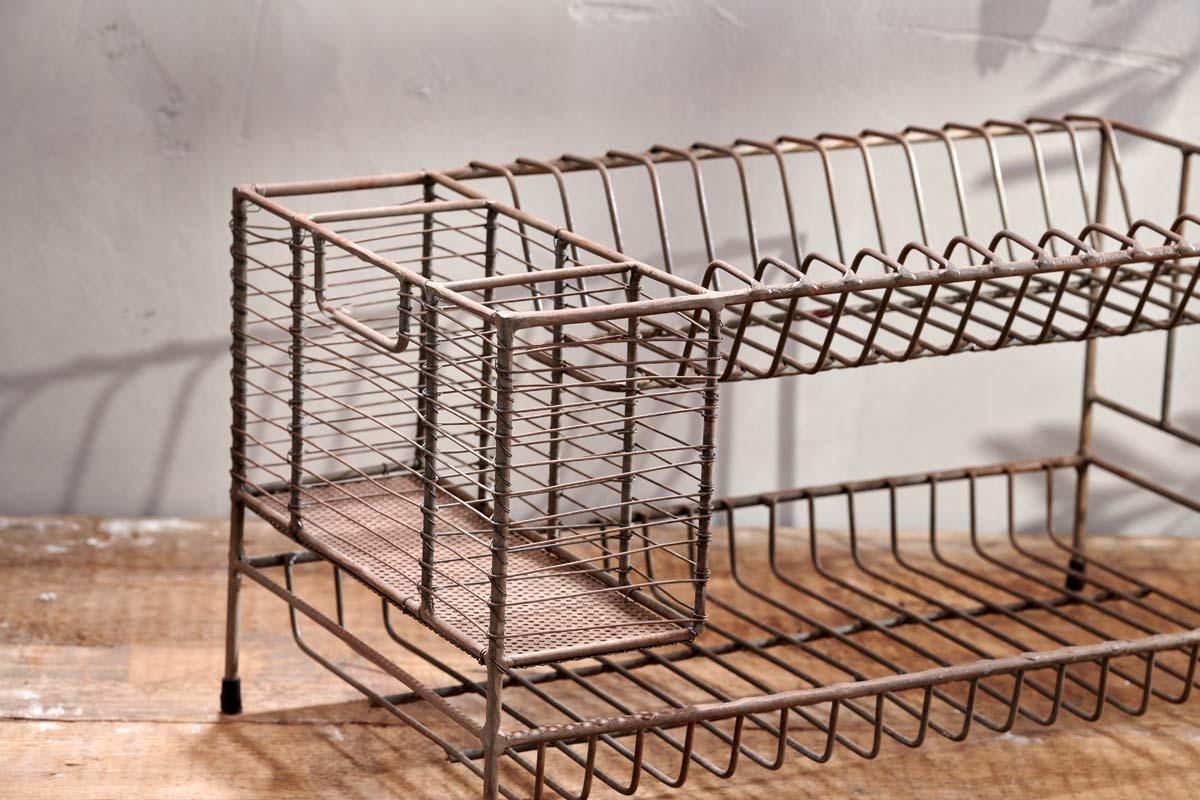The Inkollu Dish Rack by nkuku, a vintage two-tiered design with wire compartments and an aged brass finish, sits on a wooden surface against a textured light gray wall, showcasing rustic charm and visible wear.