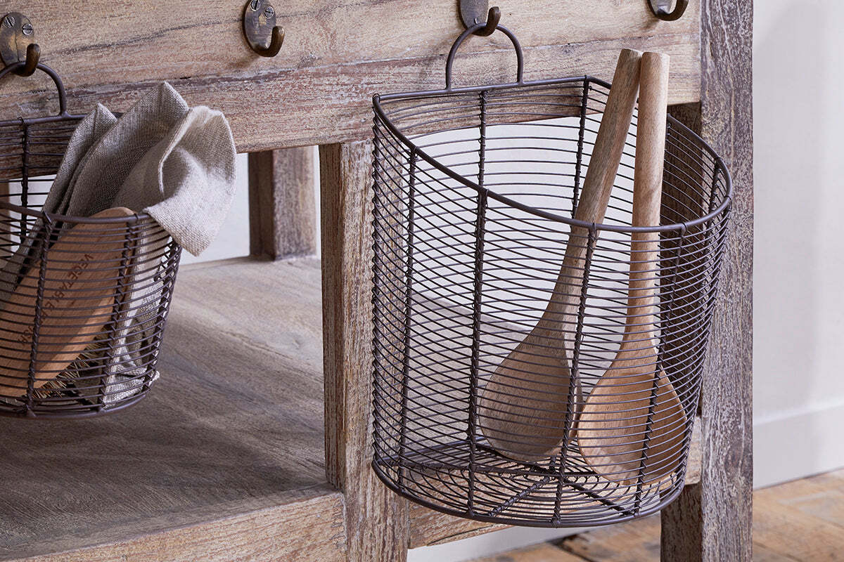 The nkuku Inkollu Wall Hung Baskets (set of 2, aged brass) hang from hooks above a rustic wooden table; one holds utensils, the other beige cloths. Stylish pantry storage on a wooden floor near a light wall.