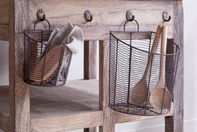 The nkuku Inkollu Wall Hung Baskets in aged brass (set of 2) hang from hooks on a rustic wooden table with a natural, weathered finish; one basket holds a beige cloth and utensil, the other two wooden spoons.