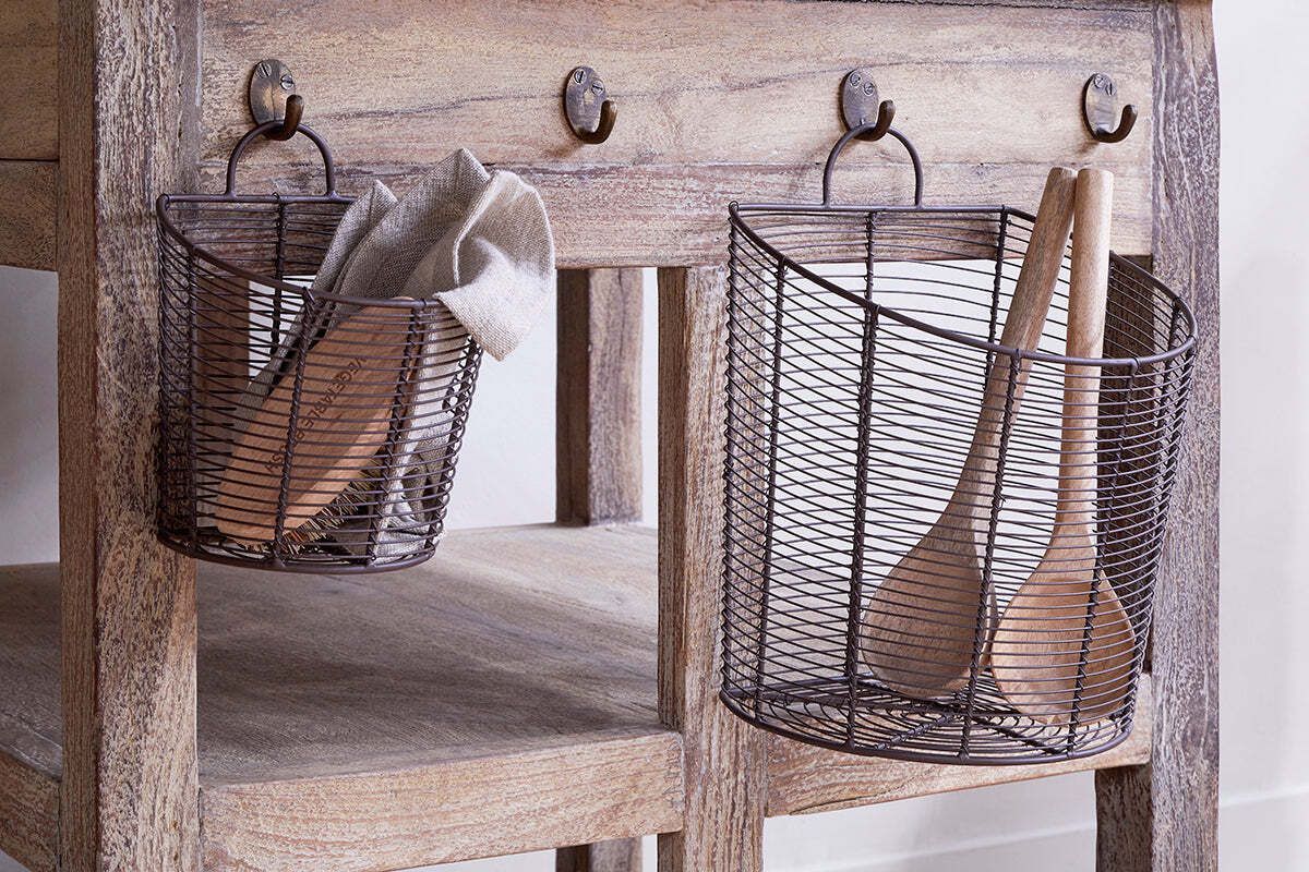 The nkuku Inkollu Wall Hung Baskets in aged brass (set of 2) hang from hooks on a rustic wooden table with a natural, weathered finish; one basket holds a beige cloth and utensil, the other two wooden spoons.