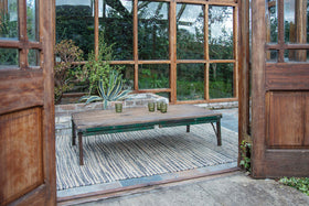 The Ishan Reclaimed Folding Dining & Coffee Table by nkuku is set with three green glasses on a striped rug. Placed before large wood-framed windows, it opens to a cozy indoor-outdoor area with green plants and wooden doors.